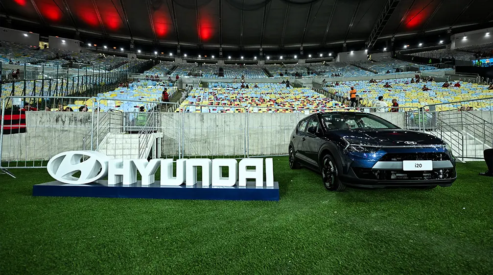 Novo Hyundai i20 ao lado de placa da Hyundai dentro de um estádio de futebol. 
