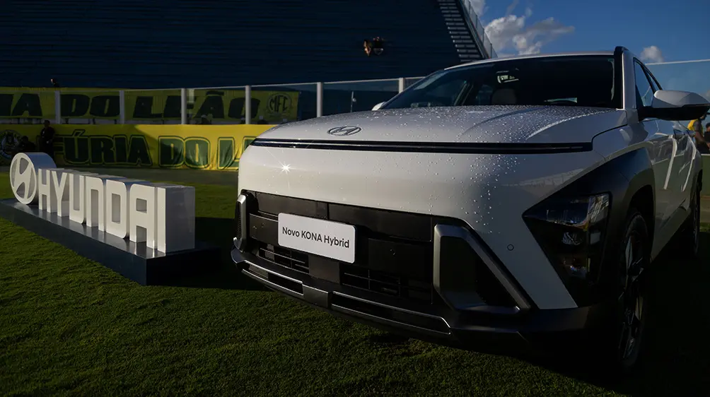 KONA Híbrido branco ao lado de placa da Hyundai dentro de um estádio de futebol. 
