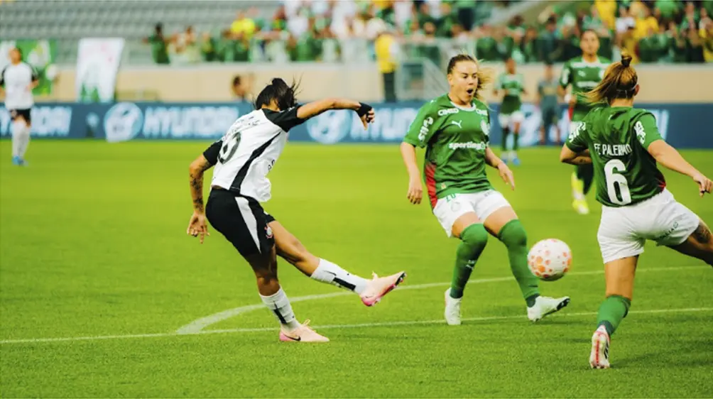 Jogadora de futebol do Corinthians chutando a bola contra o gol do Palmeiras. 