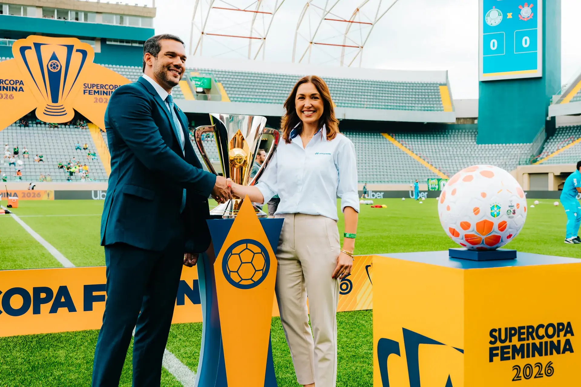 residente da CBF e representante da Hyundai apertando as mãos em um estádio de futebol com taça da SuperCopa atrás.