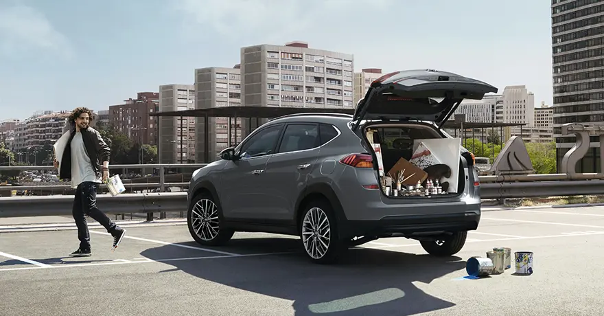 A imagem mostra o Tucson estacionado em lugar alto com o porta-malas aberto, mostrando seu espaço interno com latas de tinta, telas e pincéis, prédios da cidade ao fundo e o homem ao lado está com lata de tinta e tela nas mãos. 